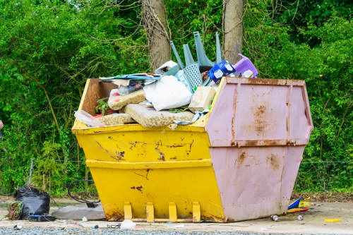 A large, open-top skip bin situated outdoors on a gravel surface, filled with mixed waste items including plastic cleaning tools, a white plastic bag, cardboard, and various household debris. The bin has a yellow front and a faded, reddish-pink back panel, with some rust and dirt visible on its surface. The environment features a backdrop of green foliage and trees, indicating a natural or semi-rural setting, with a black plastic bag and scattered small waste items on the ground nearby. The scene suggests a rubbish disposal or clearance operation, possibly associated with local rubbish removal services in Thamesmead, as managed by Office Clearance Thamesmead.