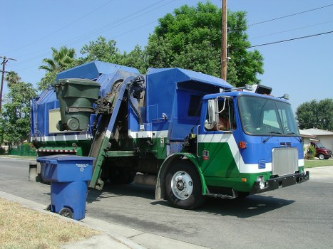 A large commercial rubbish collection truck with a predominantly blue and green colour scheme, parked on a suburban street. The truck's front cab is silver with clear glass windows and side mirrors, and it features a flat front with a rectangular shape. The rear section of the vehicle is designed for waste collection, with an open-top compactor unit that is partially raised, revealing a large, green wheeled bin positioned in front of the collection area. The bin has a slightly textured surface and a lid that is not visible. The vehicle is situated on a paved road adjacent to a grassy verge, with a driveway and residential houses partially visible in the background. Tall trees with dense green foliage line the street, and utility poles with overhead wires extend into the distance under a clear blue sky, indicating a calm day. The image is related to rubbish removal and waste management services in Thamesmead, aligning with the company Office Clearance Thamesmeadâ€™s services for waste collection in the local area.