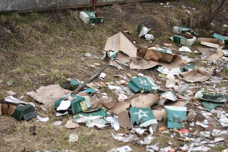 A close-up view of an unmanaged outdoor rubbish pile situated on a paved area beside a low stone wall, composed of black plastic rubbish bags, scattered paper waste, and various discarded items. Promi